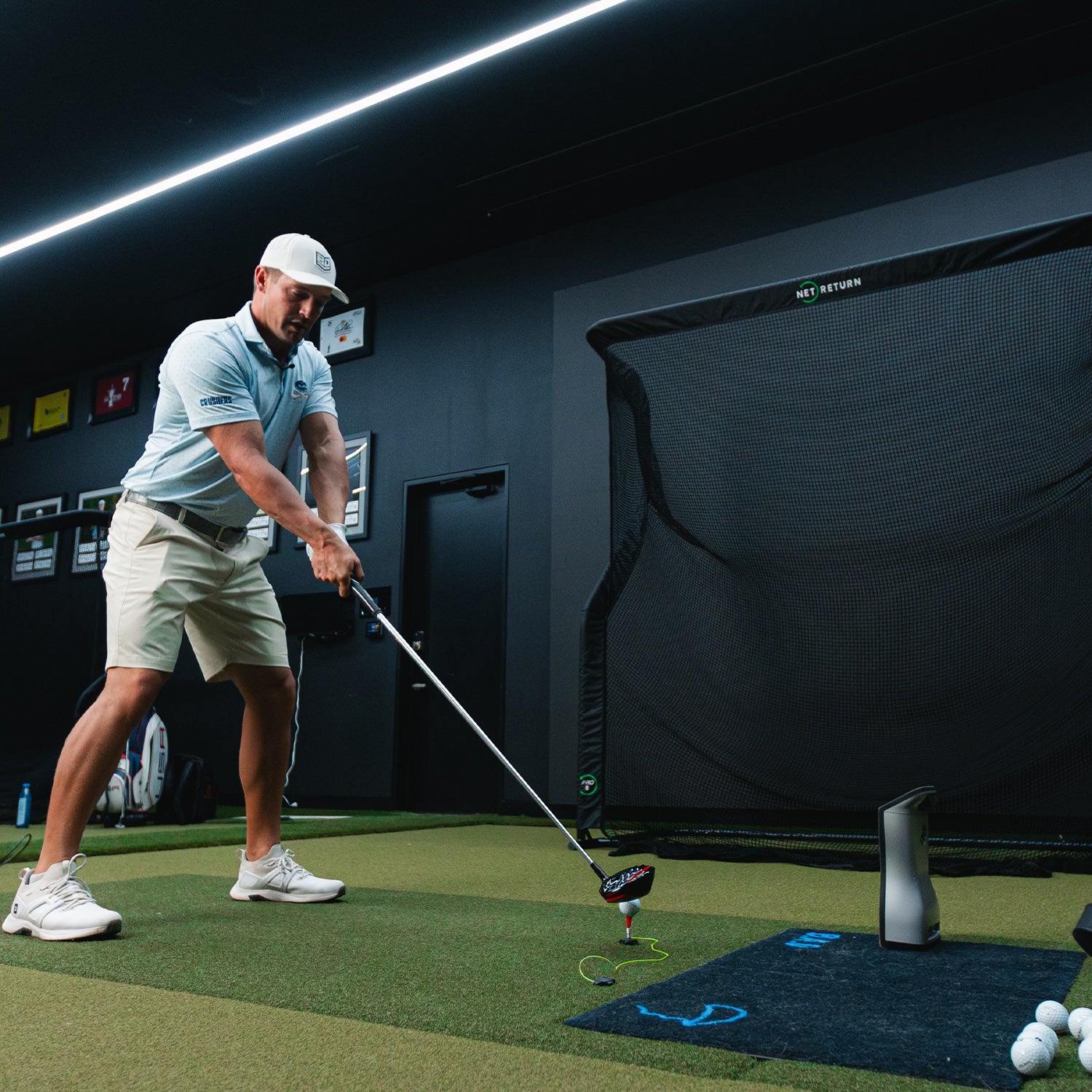 A person in a golf simulator room prepares to hit a golf ball with a driver club. Golf balls are visible on the floor, and a net for practice is in the background.