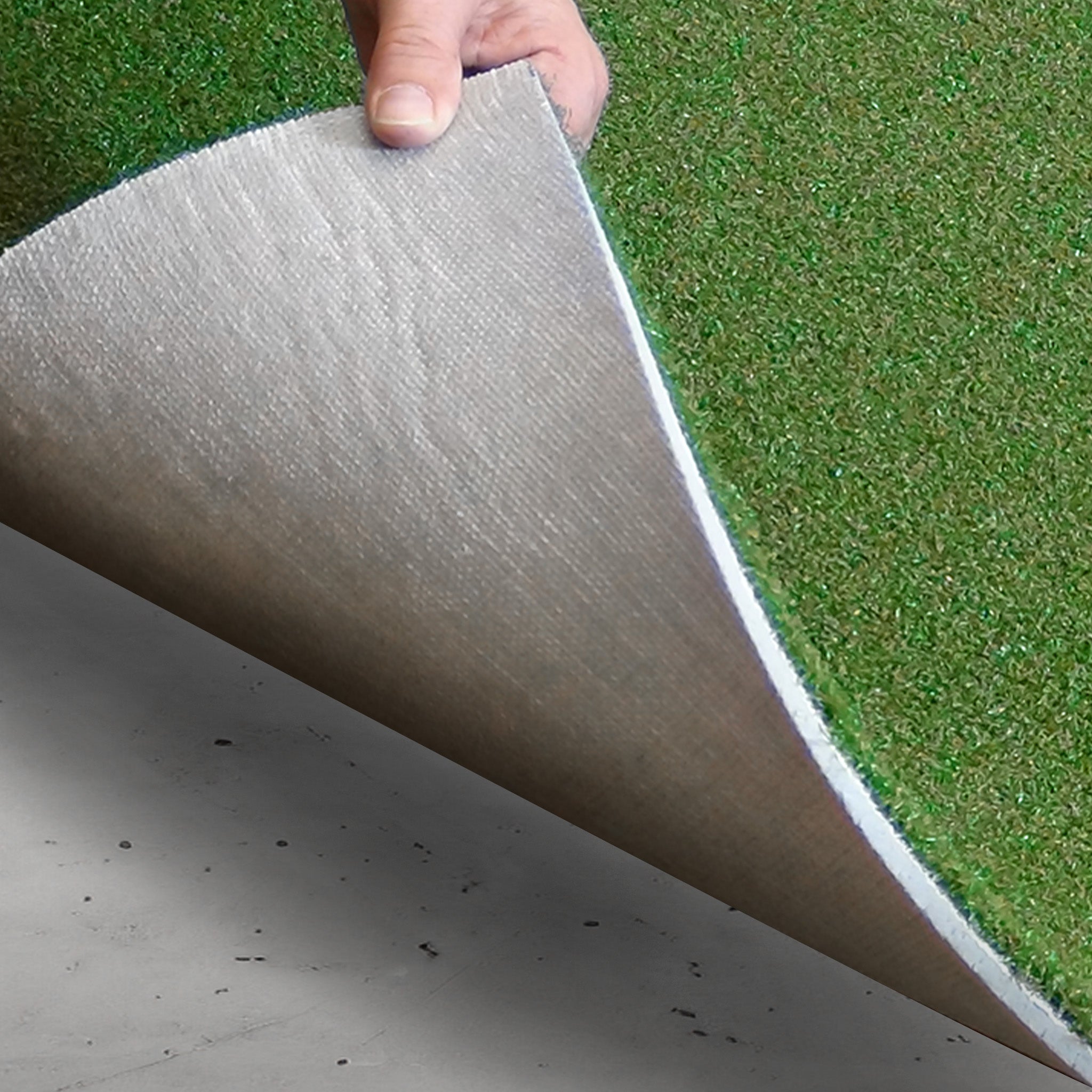 A hand lifts the edge of a piece of artificial turf, showing the textured backing against a concrete surface.